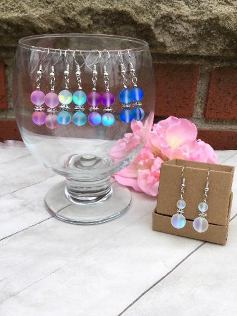 May include: A set of iridescent earrings with silver hooks and two round beads. The earrings are displayed in a glass bowl on a white surface.
