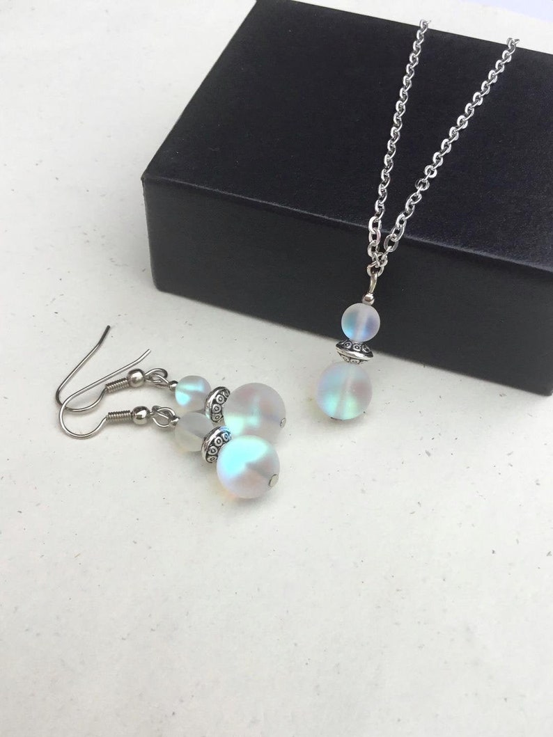 May include: A set of iridescent white opal jewelry with a silver chain necklace and matching earrings. The necklace and earrings feature a round opal bead with a silver accent.