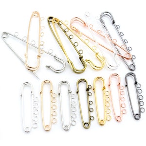 May include: A set of 12 safety pin brooches in various finishes, including silver, gold, rose gold, and black. Each pin has a series of jump rings for attaching charms or other embellishments.