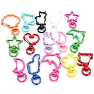 May include: Assortment of colorful metal keychains with various shapes, including a star, heart, cat, moon, and dolphin. Each keychain has a swivel clasp and a spring-loaded gate.