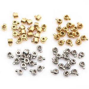 May include: A selection of small metal cord locks in gold and silver. The gold ones are cylindrical with a small hole, the silver ones are round with a side opening. These are likely used for adjusting drawstrings or cords, commonly found on clothing.