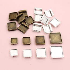 May include: A collection of square metal jewelry findings in various sizes and finishes. The findings are in antique bronze and silver tones, with some having a bezel design. The items are scattered on a pink background.
