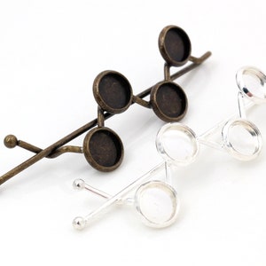 Peut inclure: Deux pinces à cheveux avec des cabochons ronds en verre. La pince à gauche est en bronze avec quatre cabochons. La pince à droite est en argent avec trois cabochons.