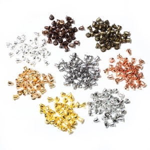 Può includere: Assortimento di piccole chiusure per orecchini in metallo di vari colori, tra cui argento, oro, oro rosa e bronzo. Le chiusure sono disposte in pile su una superficie bianca.