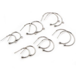 Pode incluir: Vários brincos de argola prateados com uma pequena bola na parte inferior e um pino para perfurar. Os brincos estão dispostos sobre um fundo branco. Os brincos têm um design simples e minimalista.