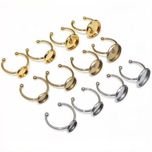 May include: A collection of twelve adjustable ring blanks featuring circular bezel settings. The set includes six gold-toned metal rings and six silver-toned metal rings, each with an open band design and a smooth, flat circular cup for mounting.