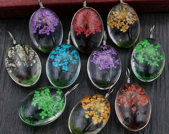 Op de afbeelding: Een verzameling ovale hangers met gedroogde bloemen ingekapseld in heldere hars. Elke hanger toont bloemen in verschillende kleuren, waaronder paars, rood, geel en groen, met een zilverkleurige draadlus om op te hangen. De hangers worden gepresenteerd op een donkere ondergrond.