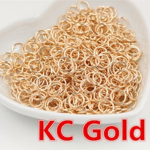 Puede incluir: Una gran cantidad de anillos de salto dorados en un cuenco blanco en forma de corazón. El cuenco está sobre una superficie blanca. El texto "KC Gold" está impreso debajo del cuenco.