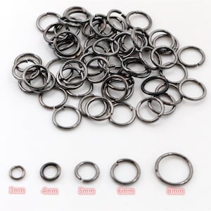 Puede incluir: Un montón grande de anillos de salto de metal negro en varios tamaños, que van de 3 mm a 8 mm. Los anillos están dispuestos en un patrón aleatorio sobre una superficie blanca.