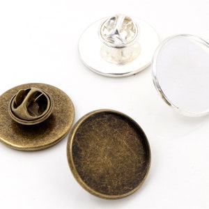 Op de afbeelding: Twee ronde metalen bases voor het maken van aangepaste pinnen. Eén basis is zilver en de andere is antiek messing. Beide bases hebben een pin achterkant.