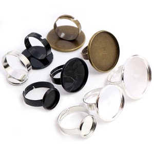 May include: Assortment of adjustable ring blanks with round glass cabochon settings. The settings are in various sizes and colors, including black, silver, and bronze.