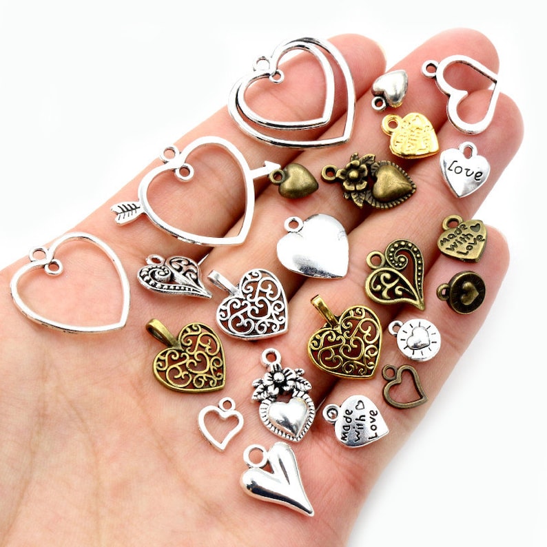 K&ouml;nnte beinhalten: Sortiment an silbernen und goldfarbenen herzf&ouml;rmigen Charms mit verschiedenen Designs, darunter Filigran, durchbrochenes Muster und gravierte Botschaften wie "love" und "made with love".