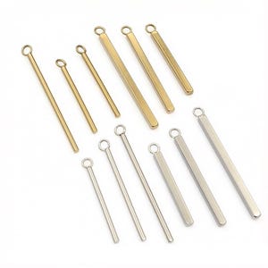 May include: A collection of twelve metal bar charms arranged in two rows, featuring six gold-toned and six silver-toned pieces. Each charm consists of a long, slender rectangular bar with a small circular jump ring attached to the top end.