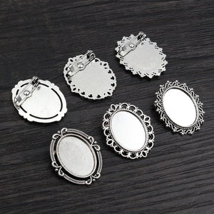Puede incluir: Seis ajustes de broche de camafeo de metal plateado con diseños de filigrana ornamentados. Los ajustes son de forma ovalada y tienen una superficie lisa y plana para agregar un camafeo u otro adorno.
