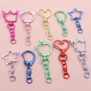 Könnte beinhalten: Eine Sammlung bunter Metall-Schlüsselanhänger in verschiedenen Formen, darunter Sterne, Kronen, Herzen und Äpfel. Die Schlüsselanhänger sind in den Farbtönen Rosa, Gold, Grün, Blau und Rot gehalten, mit einem Karabinerverschluss und einer Kette.