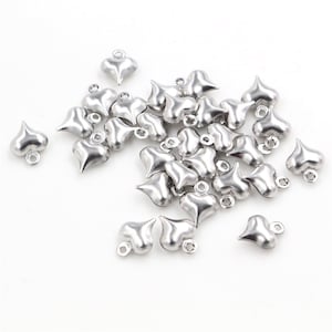May include: A pile of silver heart-shaped charms with a loop at the top. The charms are scattered on a white background.