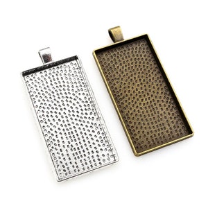 Könnte beinhalten: Zwei rechteckige Metall-Anhänger-Tabletts, eines silberfarben und eines bronzefarben, mit einer strukturierten Oberfläche. Beide Tabletts haben oben eine Öse zum Aufhängen.