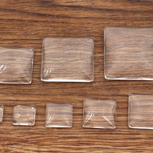 Könnte beinhalten: Sortiment aus acht quadratischen Glascabochons in verschiedenen Größen, auf einer Holzoberfläche angeordnet.