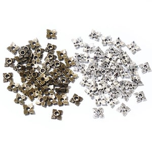 Op de afbeelding: Een grote stapel zilveren en bronzen metalen bloemvormige kralen. De kralen zijn verspreid over een wit oppervlak.