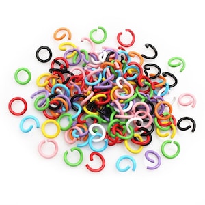 Puede incluir: Un conjunto de anillos abiertos de colores variados: rojo, naranja, amarillo, verde, azul, morado, rosa, negro y blanco. Estos pequeños anillos en forma de C están dispersos sobre una superficie blanca, ideales para la creación de joyas y manualidades.