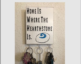May include: A white wooden sign with black text that reads "Home is where the Hearthstone is."  The sign has a blue Hearthstone logo painted on it and is mounted on a wall with four hooks holding a set of keys.