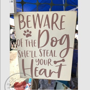 May include: A white wooden sign with brown text that reads "Beware of the Dog She'll Steal Your Heart" with a paw print and a bone graphic.
