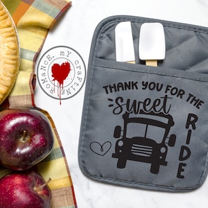 May include: Grey oven mitt with a pocket for utensils. The mitt has a black graphic of a school bus with the text "Thank you for the sweet ride".