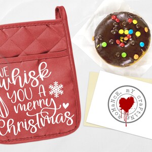 Pode incluir: Luva de forno acolchoada vermelha com o texto "We whisk you a merry Christmas" em letras brancas. Um batedor branco está incluído no texto. Um floco de neve e um pequeno coração também estão incluídos no texto.