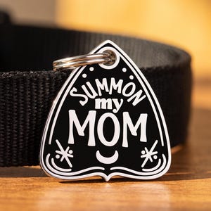 May include: A black and white pet ID tag shaped like a planchette, featuring the text "SUMMON my MOM" in a bold font. The tag includes a small crescent moon and star-like symbols, attached to a black nylon collar with a silver metal ring.