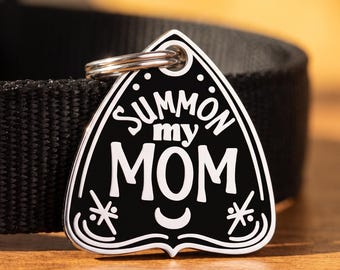 May include: A black and white pet ID tag shaped like a planchette, featuring the text "SUMMON my MOM" in a bold font. The tag includes a small crescent moon and star-like symbols, attached to a black nylon collar with a silver metal ring.