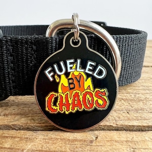 May include: A black pet tag with the words "Fueled By Chaos" in white and red lettering, with a yellow and orange flame design. The tag is attached to a black pet collar with a silver metal ring.