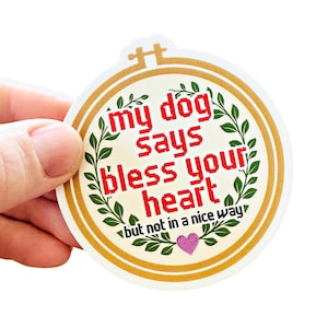 May include: A circular sticker with a gold border. The text "my dog says bless your heart but not in a nice way" is in a red pixelated font. Green leaf accents and a pink heart are also featured.