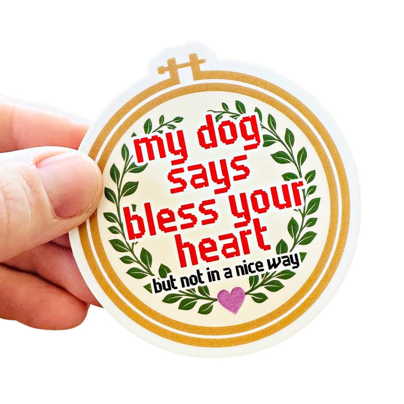 May include: A circular sticker with a gold border. The text "my dog says bless your heart but not in a nice way" is in a red pixelated font. Green leaf accents and a pink heart are also featured.