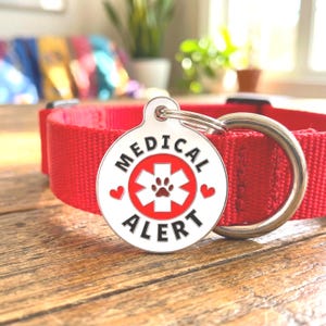 Puede incluir: Un collar rojo para perro con una etiqueta metálica plateada. La etiqueta tiene las palabras "MEDICAL ALERT" en letras negras alrededor de un símbolo médico rojo con una huella de pata. Dos corazones rojos flanquean el símbolo.