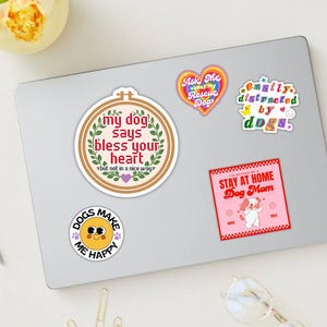 May include: A selection of dog-themed stickers on a laptop. The stickers display phrases such as "My dog says bless your heart," "Easily distracted by dogs," and "Stay at home Dog Mom." Other stickers include a heart and a smiling face.
