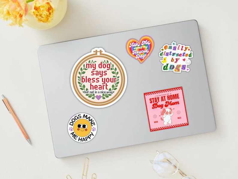 May include: A selection of dog-themed stickers on a laptop. The stickers display phrases such as "My dog says bless your heart," "Easily distracted by dogs," and "Stay at home Dog Mom." Other stickers include a heart and a smiling face.