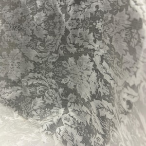 May include: A close-up shot of a sheer fabric with a repeating floral pattern in grey and white. The design features stylised leaves and flowers, creating a delicate texture. The fabric appears lightweight and slightly translucent.