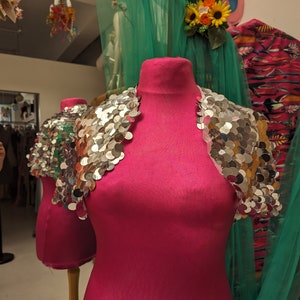 May include: A pink dress form with a silver sequined bolero on top. The bolero has a scalloped edge and is made of small, round sequins.
