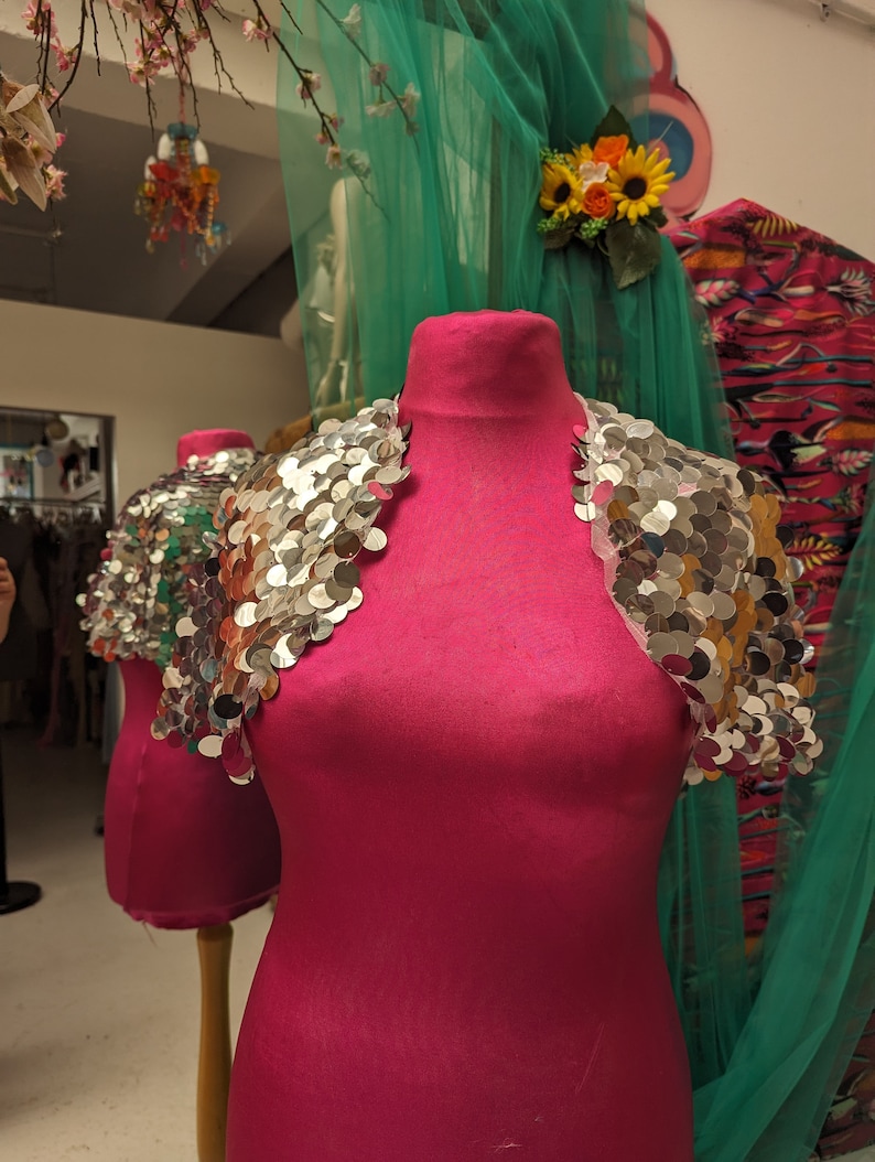 May include: A pink dress form with a silver sequined bolero on top. The bolero has a scalloped edge and is made of small, round sequins.