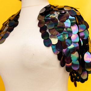 May include: A black mesh shrug with iridescent sequins in a circular pattern. The sequins are arranged in a cascading design, creating a dramatic and eye-catching look.