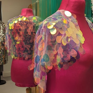 May include: Two iridescent sequin fabric shoulder wraps on pink mannequins. The sequins are round and have a holographic finish.