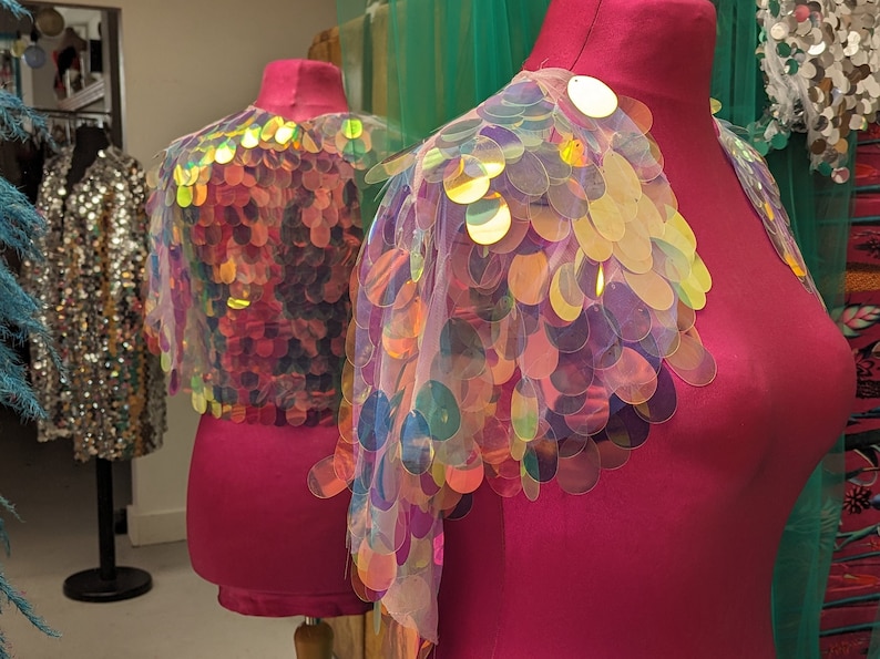 May include: Two iridescent sequin fabric shoulder wraps on pink mannequins. The sequins are round and have a holographic finish.