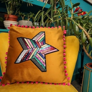 May include: Mustard-colored velvet throw pillow with a large star design. The star is made of colorful sequins and embroidery. The pillow has a pink pom-pom trim and is placed on a yellow chair.