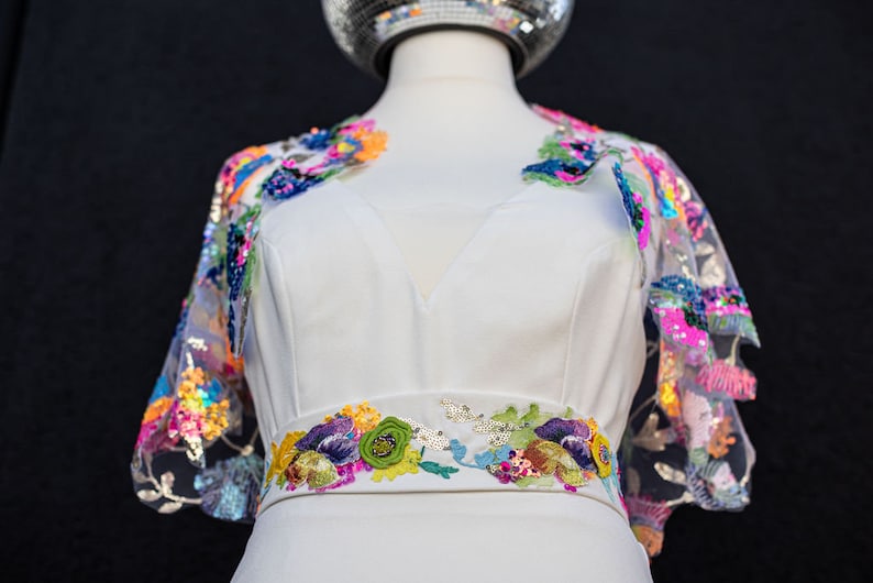 May include: A white dress with a colourful floral embroidered belt and matching floral embroidered sleeves. The sleeves are sheer and have sequins.