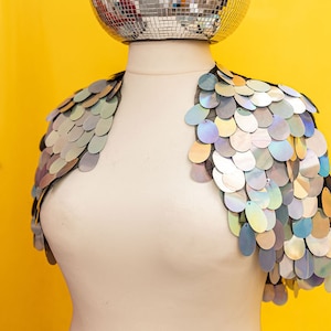 May include: A silver sequined shrug with iridescent sequins. The shrug is on a white mannequin against a yellow background.