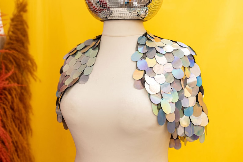 May include: A silver sequined shrug with iridescent sequins. The shrug is on a white mannequin against a yellow background.
