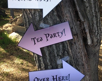 May include: Three arrow signs taped to a tree trunk. The top sign is white with black text that reads "WRONG WAY!" The middle sign is purple with black text that reads "Tea Party!" The bottom sign is blue with black text that reads "Over Here!"