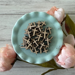 May include: A collection of small, wooden crosses arranged on a light blue, ruffled-edge plate. The crosses are a natural wood color. The plate is surrounded by pink flowers and green leaves, set against a white-washed wooden background.
