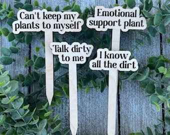 Könnte beinhalten: Vier hölzerne Pflanzenmarker mit humorvollen Sätzen in schwarzem Text: "Can't keep my plants to myself", "Emotional support plant", "Talk dirty to me" und "I know all the dirt". Die Marker sind vor einem Hintergrund aus Grün und einer verwitterten Holzoberfläche platziert.