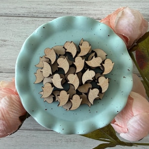 May include: A collection of small, wooden ghost-shaped cutouts arranged on a light blue, scalloped-edge plate. The cutouts are a light brown color. Pink flowers are in the background.